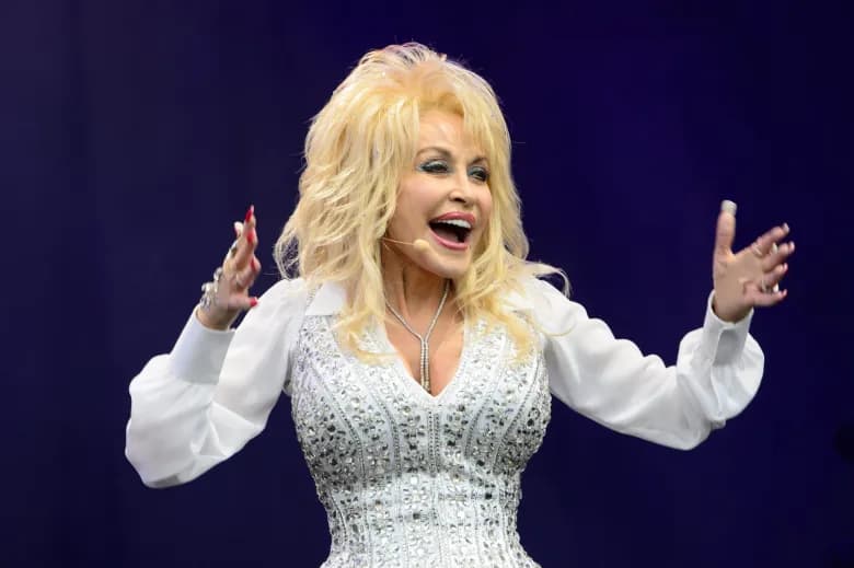 Dolly Parton performing on stage in a white embellished outfit, arms raised, wearing a headset microphone
